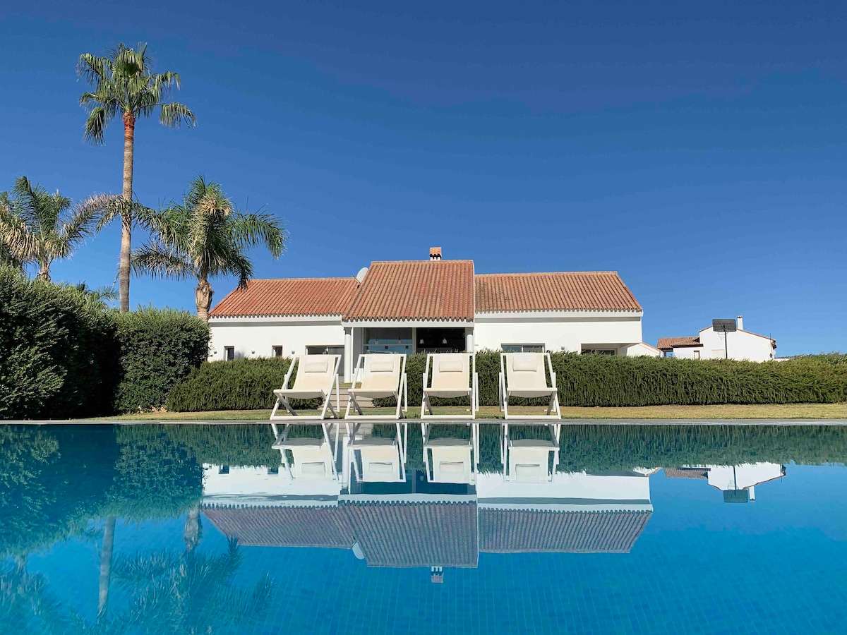 A serene pool area features four sun loungers positioned beside a clear, inviting pool. The reflection of the villa with a red-tiled roof and surrounding greenery is visible on the water's surface under a clear blue sky.