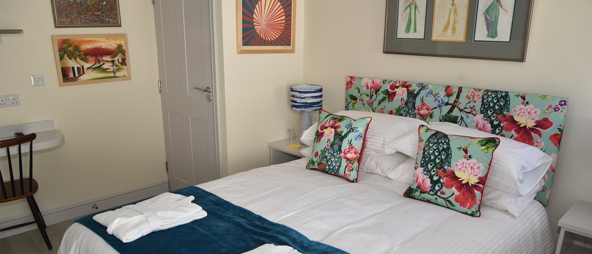 A bright bedroom features a spacious bed with floral-patterned headboard and decorative cushions. Artwork adorns the walls, adding color to the space. A wooden chair and desk are positioned nearby, alongside a bedside lamp, creating a cozy atmosphere.