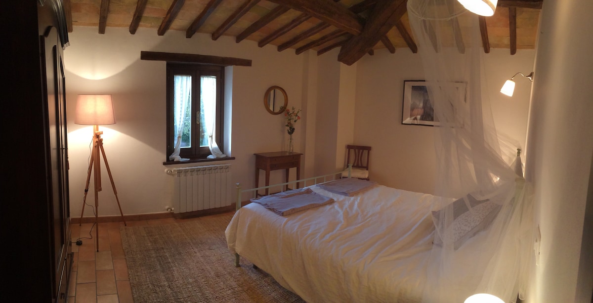 A spacious bedroom features a comfortable bed covered with soft linens. Natural light enters through a window, complemented by a decorative mirror and a small wooden table. A floor lamp and a light fixture provide soft illumination, while the ceiling showcases wooden beams.