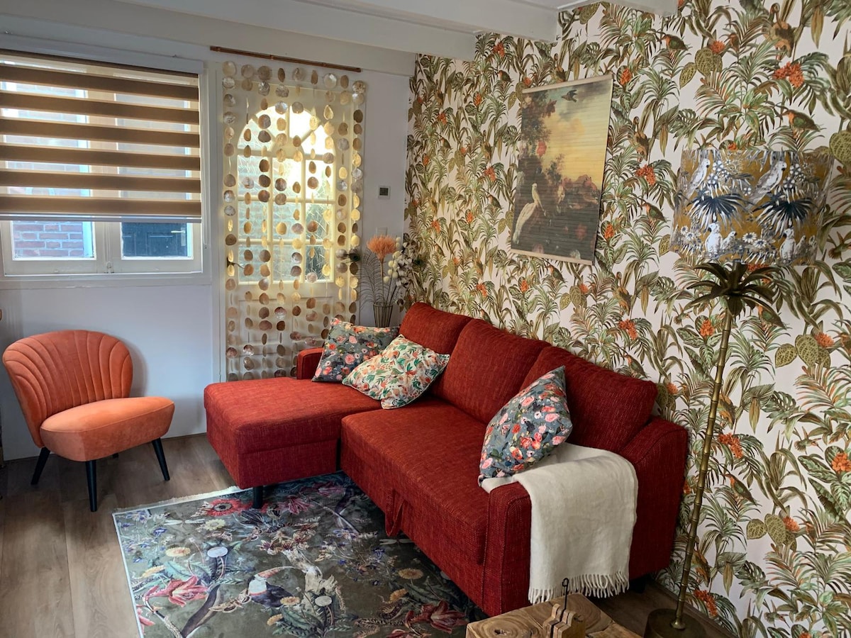 A cozy living area features a red sectional sofa adorned with patterned cushions, complemented by an orange accent chair. A floral wallpaper creates a vibrant backdrop, while a decorative lamp stands beside the sofa. A patterned rug adds warmth to the space.