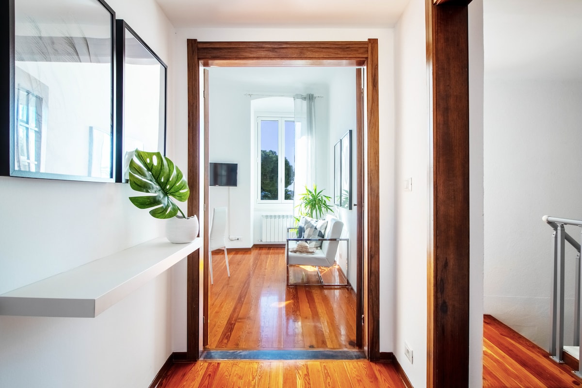 A hallway is visible, showcasing wooden flooring and white walls. A small shelf holds a green plant, while light streams through a window at the end. The adjacent room features a comfortable seating area with a plant and an entertainment unit.