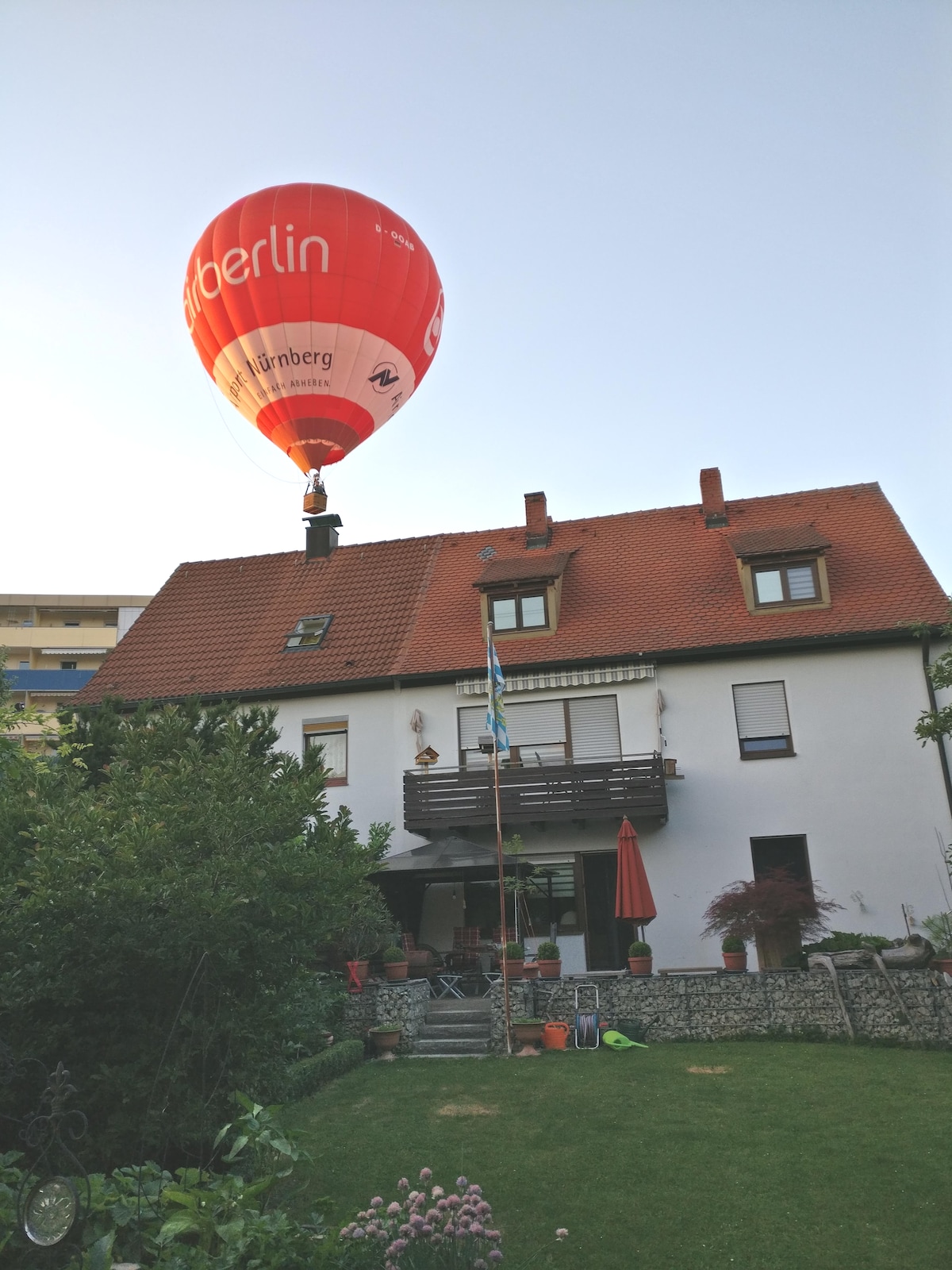 A red hot air balloon is seen rising above a multi-story house with a green garden below. The house features a combination of white and brick exterior, with a balcony and several windows. A patio area with a red umbrella is visible in the garden.
