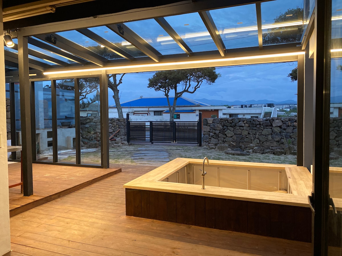 An outdoor deck area is seen, featuring a small white hot tub and wooden flooring. Large glass panels provide a view of the surrounding landscape, while overhead lighting enhances the space's openness. A stone wall borders the property, adding a rustic touch.