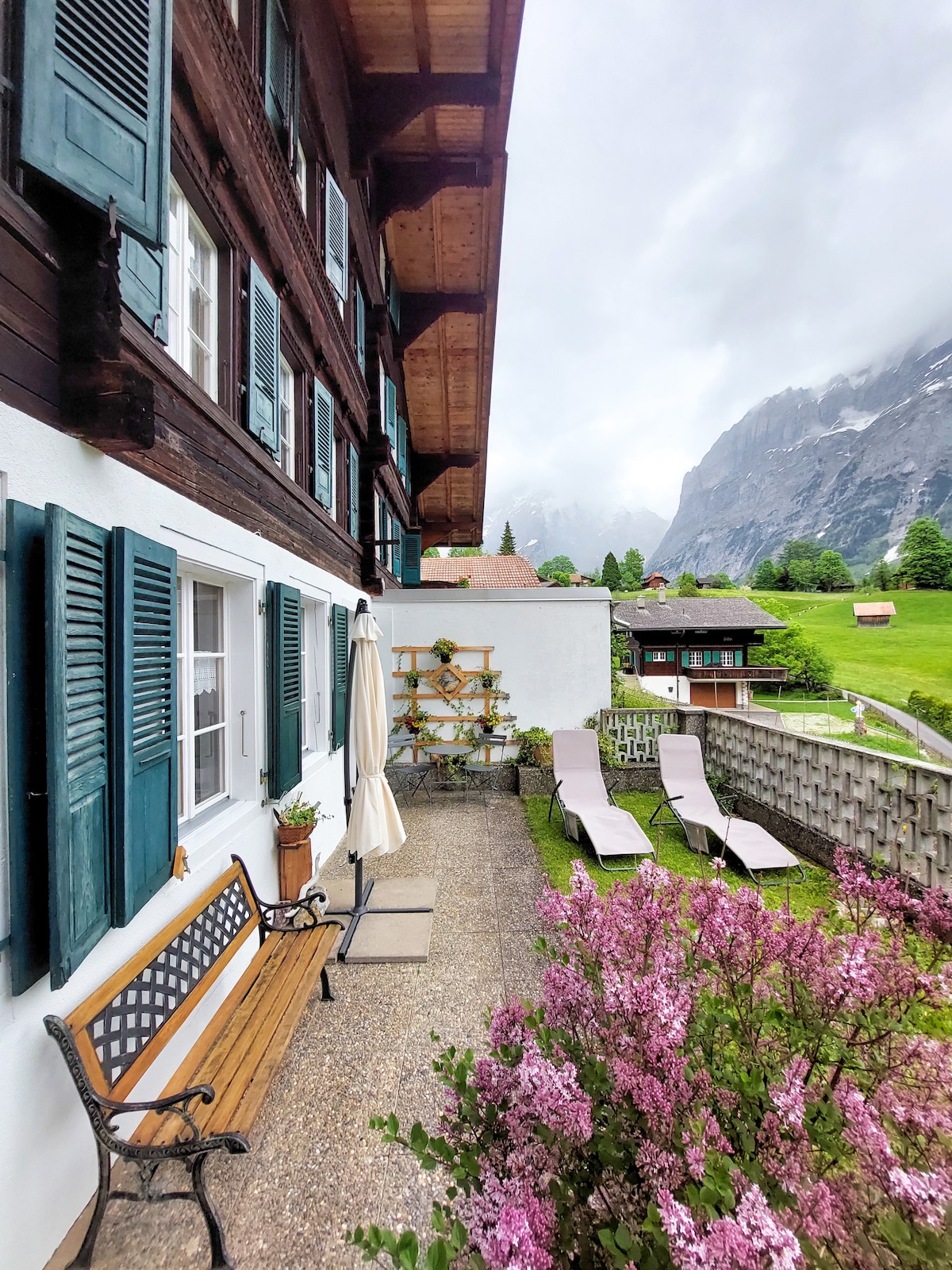 A charming outdoor seating area features a wooden bench and sun loungers set against a backdrop of greenery and distant mountains. The space is enhanced by blooming lilacs and is partially shaded by an umbrella, offering a serene environment for relaxation.