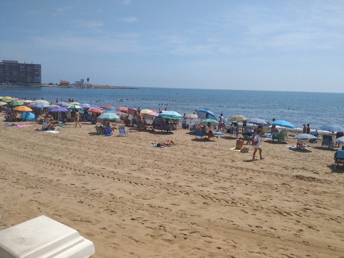 A sandy beach scene is depicted, featuring colorful umbrellas providing shade over beachgoers relaxing on lounge chairs. The calm sea extends to the horizon, with a few people swimming. In the distance, buildings are visible along the coastline.