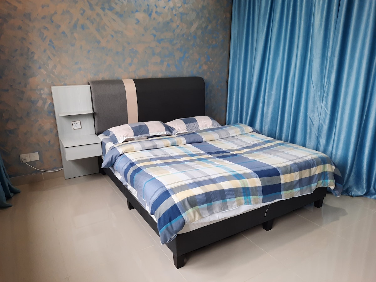 A comfortable bedroom setting features a spacious bed with a patterned blanket. Soft blue curtains frame the window, enhancing the tranquil ambiance. A bedside table stands beside the bed, offering functional space for personal items against a lightly textured wall.