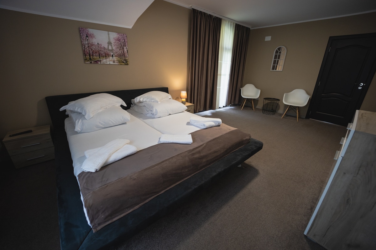 A spacious bedroom features a large bed with white linens and dark brown accents, complemented by two chairs positioned near a window. Soft lighting creates a calming ambiance, while a piece of artwork depicting a serene landscape hangs above the bed.