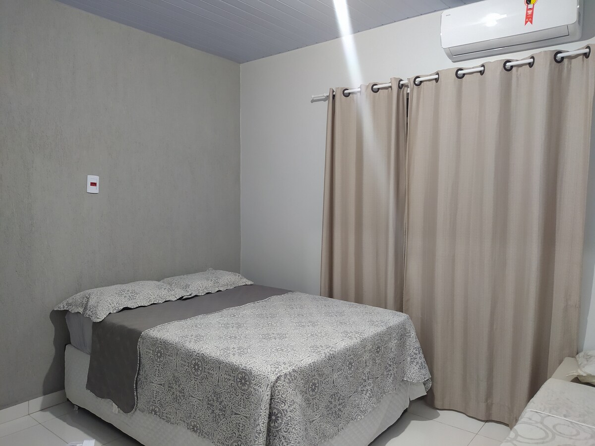 A sleeping area features a spacious bed with a light-colored bedspread, complemented by a darker blanket. Neutral-toned curtains frame a window, with an air conditioning unit positioned above. The room is designed with a minimalistic style and a calm color palette.