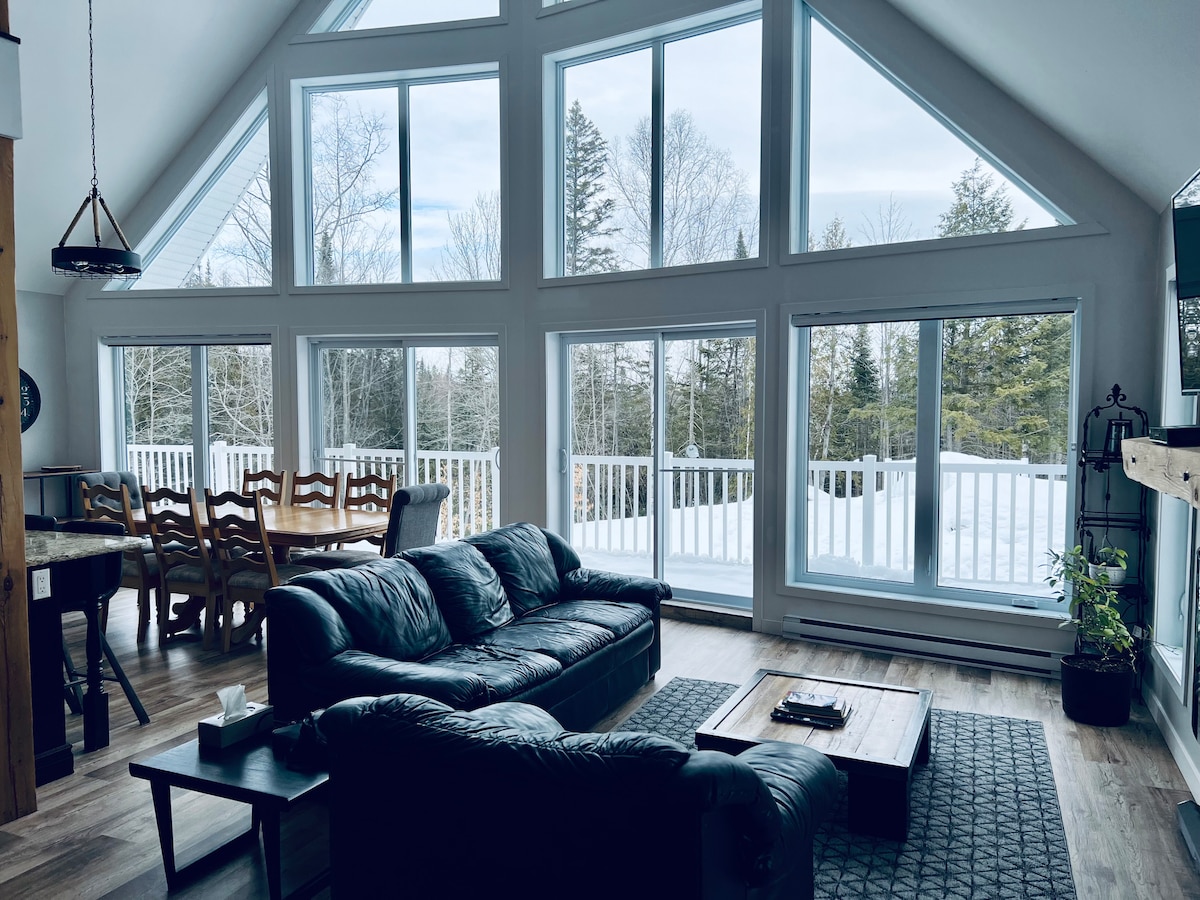 A welcoming living area features a large sectional sofa facing expansive windows that allow natural light to fill the space. A coffee table is positioned centrally on a textured area rug. A dining table is visible in the background, surrounded by wooden chairs.