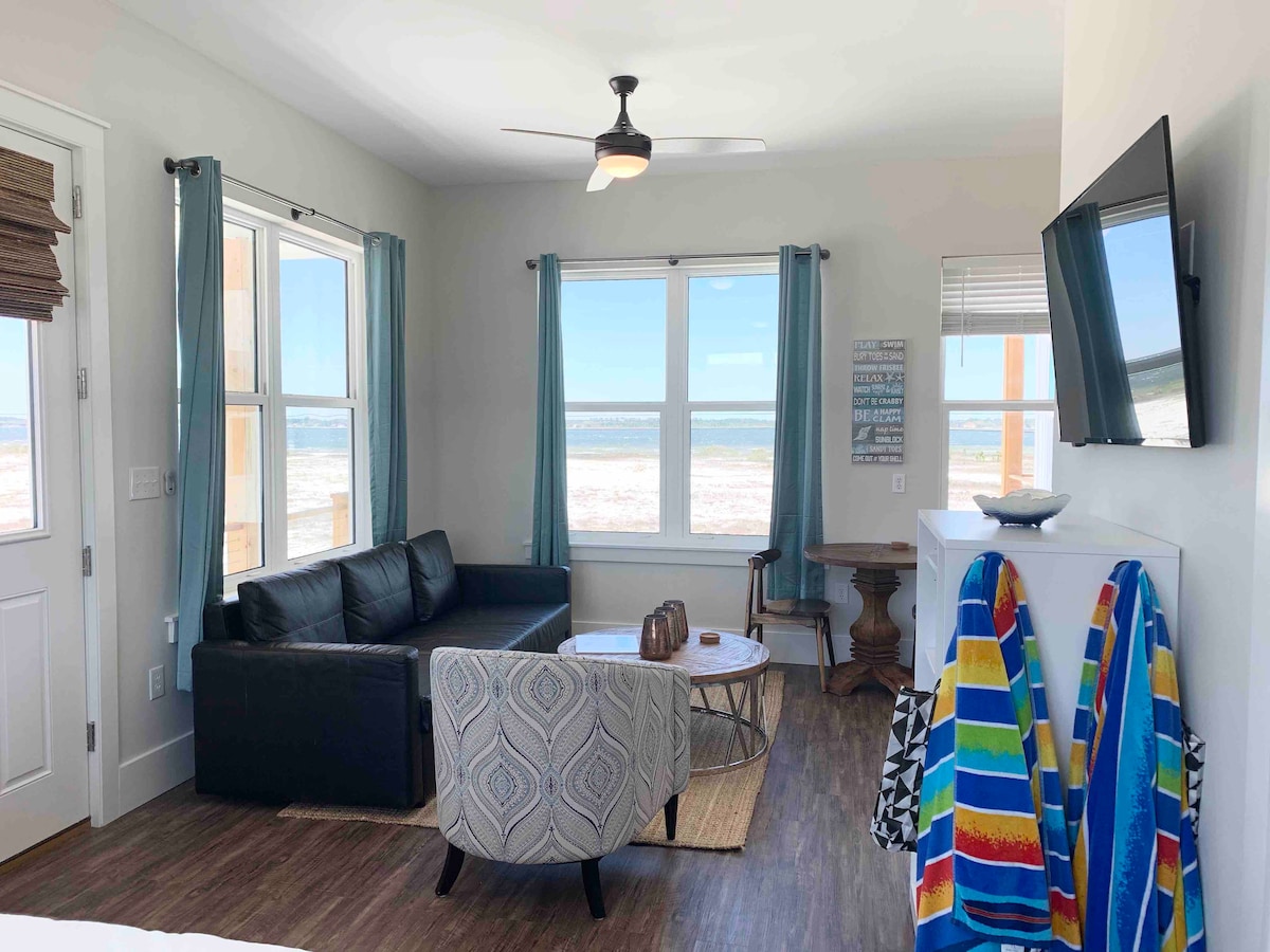 A brightly lit living area features large windows offering views of the beach. A dark couch is paired with a round coffee table and a decorative rug. Colorful beach towels hang nearby, adding a vibrant touch to the space.