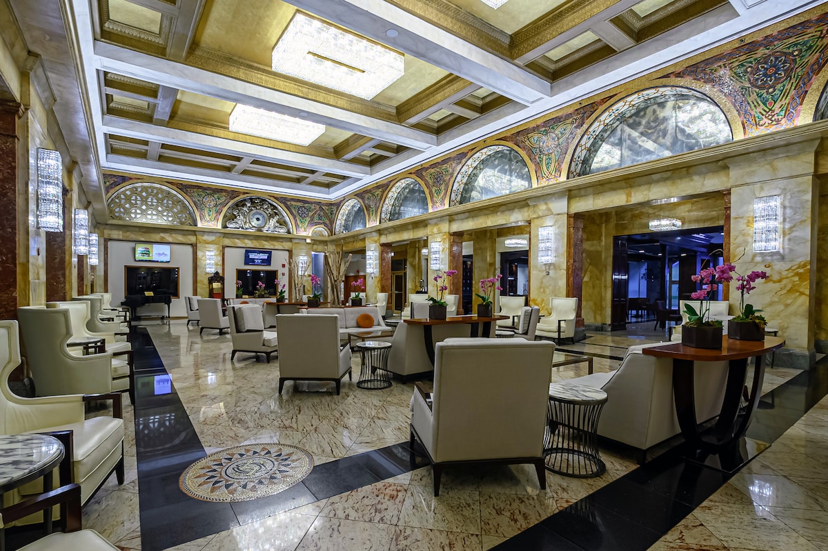 A grand lobby is highlighted by ornate ceilings and detailed arches, creating an upscale ambiance. Elegant seating arrangements consist of plush chairs and stylish tables, all set against polished marble floors. Decorative plants and floral displays add a touch of vibrancy to the space.