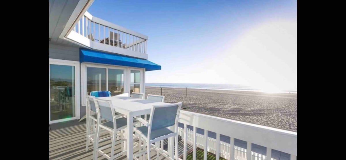 A sunlit deck features a white outdoor dining table surrounded by tall chairs, overlooking a sandy beach and ocean view. The bright blue awning adds contrast against the clear sky, while the expansive beach extends into the distance.