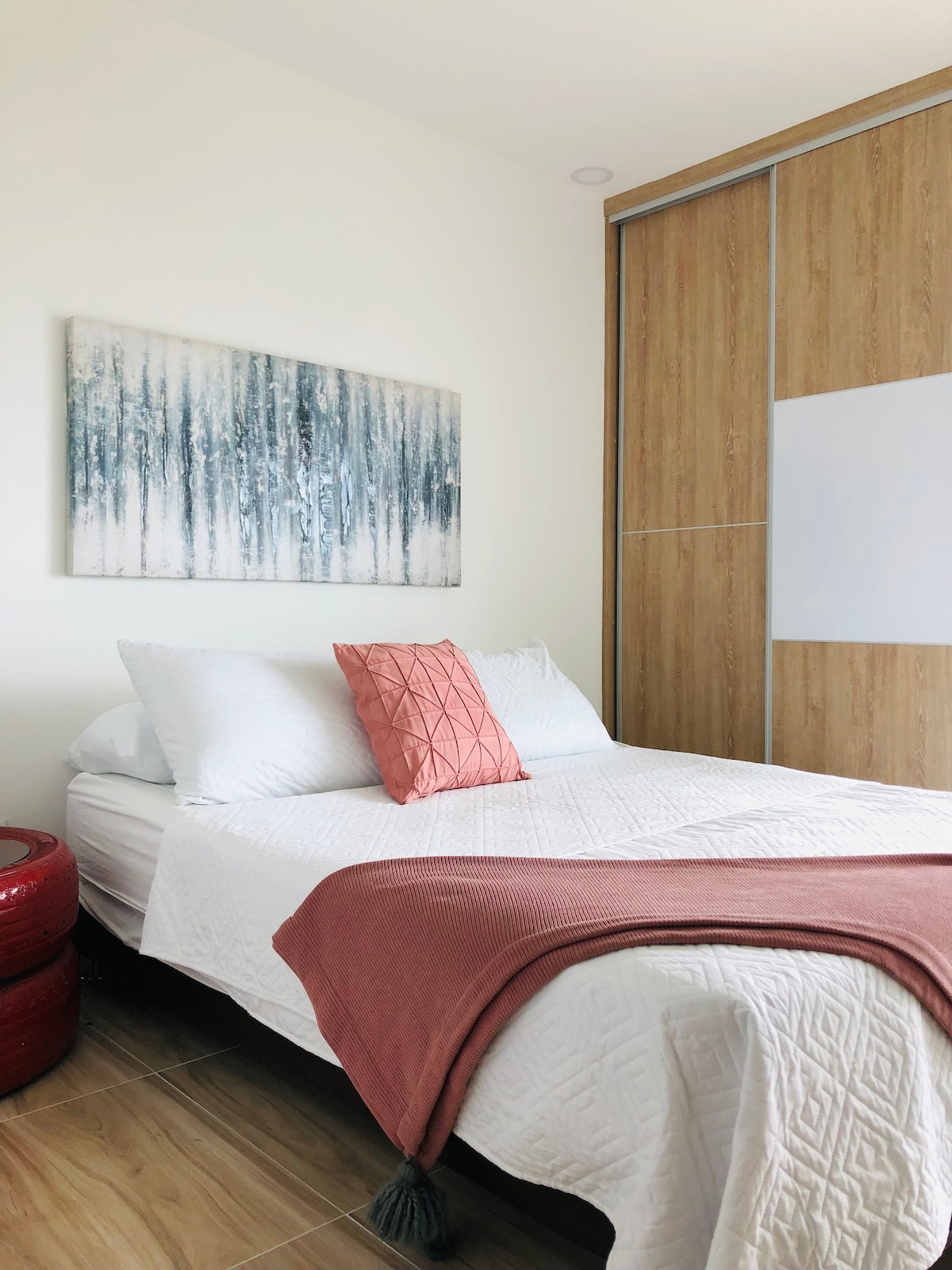 A cozy sleeping area is defined by soft white bedding and a decorative pink pillow. Light wood accents on the cabinetry complement the textured wall art. A warm-toned throw blanket drapes casually over one side of the spacious bed, contributing to a calming atmosphere.