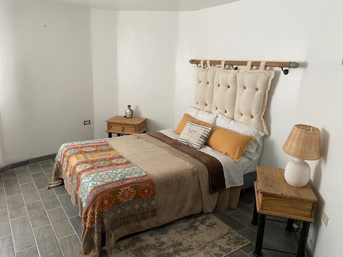 A comfortable sleeping area features a spacious bed with a decorative headboard and warm-colored bedding. Nightstands with lamps are positioned on either side, while natural light enters from a window, illuminating the textured flooring.