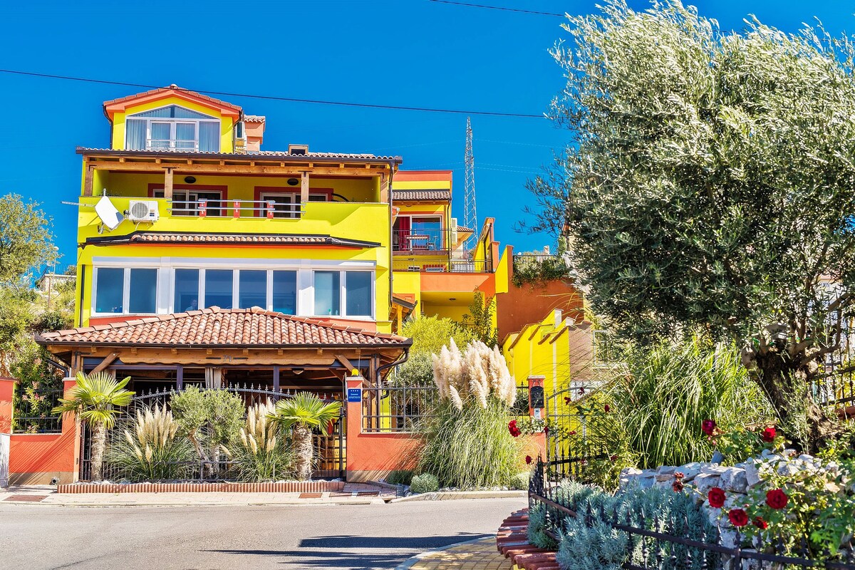 A vibrant multi-story building is surrounded by lush greenery and colorful landscaping. Large windows reflect natural light, while a tiled roof complements the bright yellow exterior. Palm trees and flowering plants enhance the inviting entryway, creating a welcoming atmosphere.