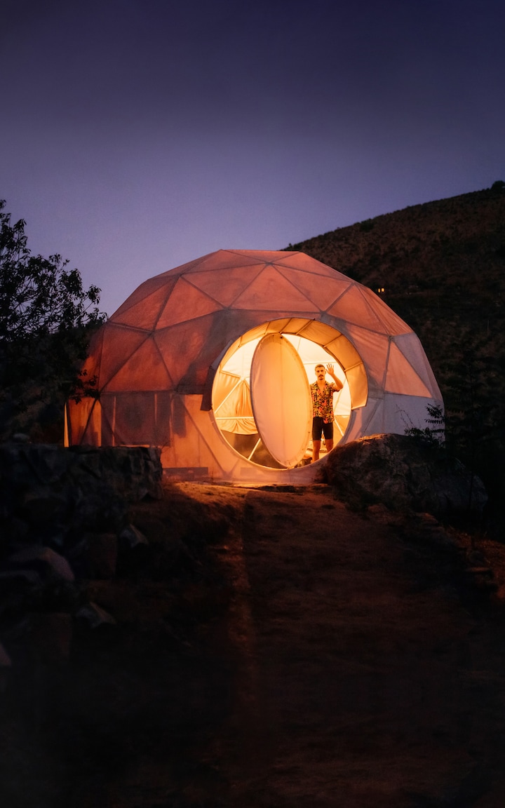 A guest stands, waving, in the doorway of a unique-looking dome house at dusk, bathed in warm, welcoming light.
