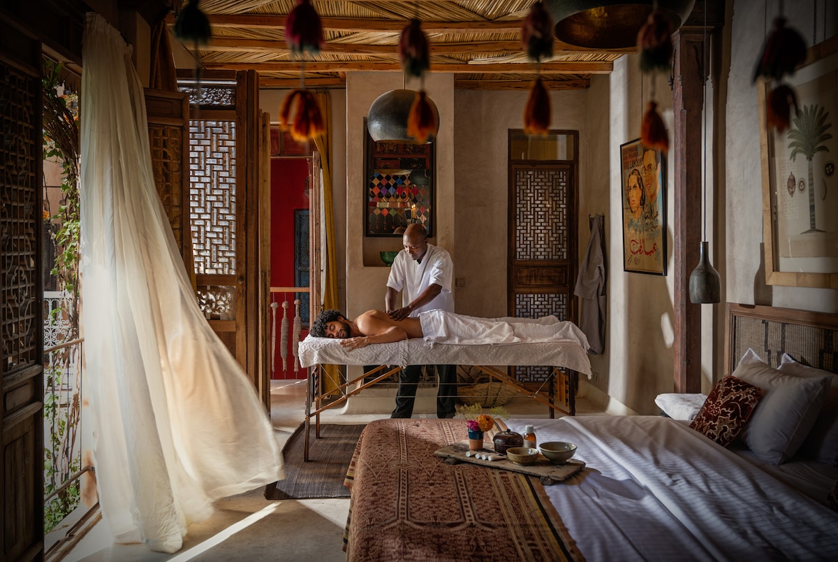 A massage room is captured, featuring a treatment table centered in a naturally lit space. The room showcases carved wooden doors and soft drapes, enhancing the serene ambiance. A low table is adorned with fragrant oils and fruits, adding to the relaxation experience offered in the room.