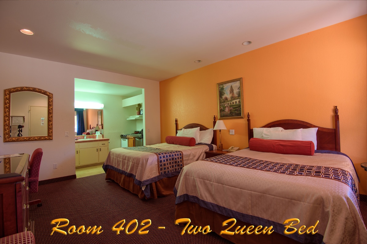 A spacious room features two queen beds with decorative covers. An orange accent wall adds warmth, while a kitchenette area is visible in the background. A small desk is placed near the entrance, and natural light is provided by a window dressed with simple curtains.