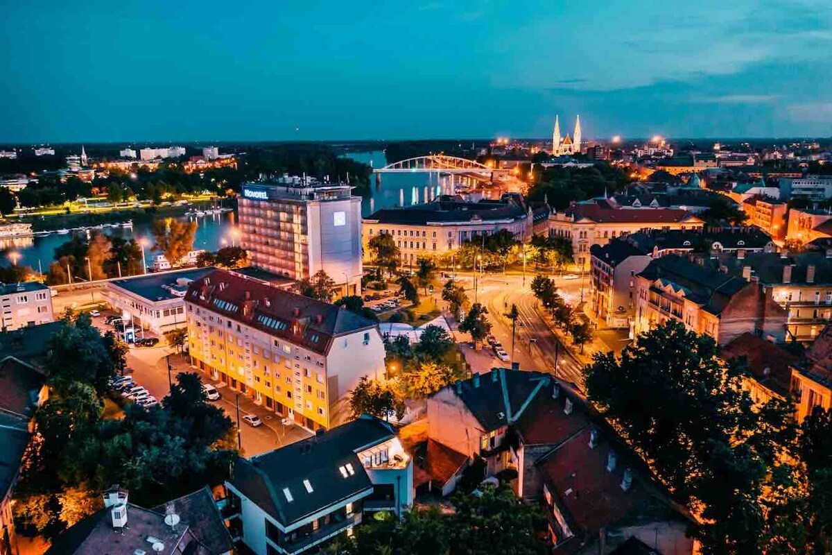 An aerial view captures a vibrant cityscape during twilight, showcasing illuminated buildings and bridges. The river reflects the evening lights, while tree-lined streets and parked cars add to the urban atmosphere. Historic and modern architecture create a diverse skyline.
