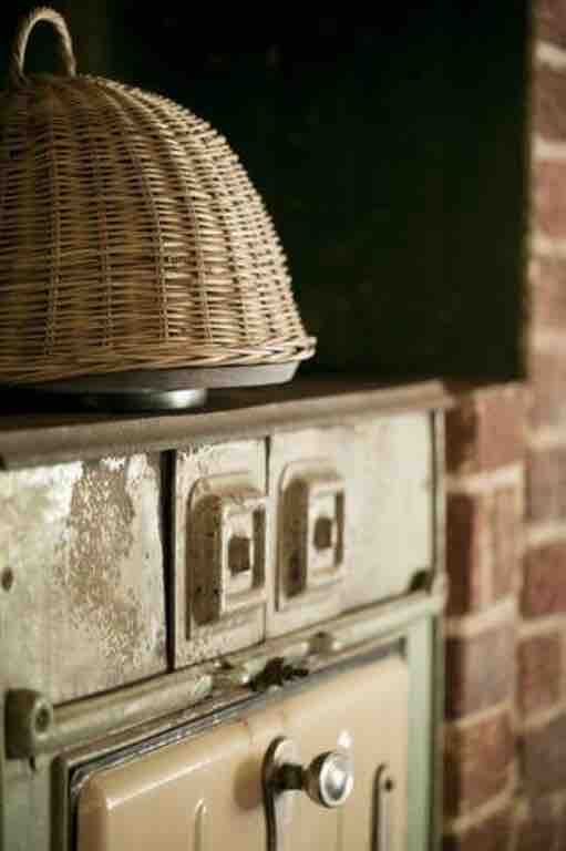 An antique stove is presented against a rustic brick wall, featuring a vintage design and subtle wear. A woven basket sits atop the stove, adding an element of texture to the scene. The overall ambiance reflects the charm of a restored 1920s cottage.