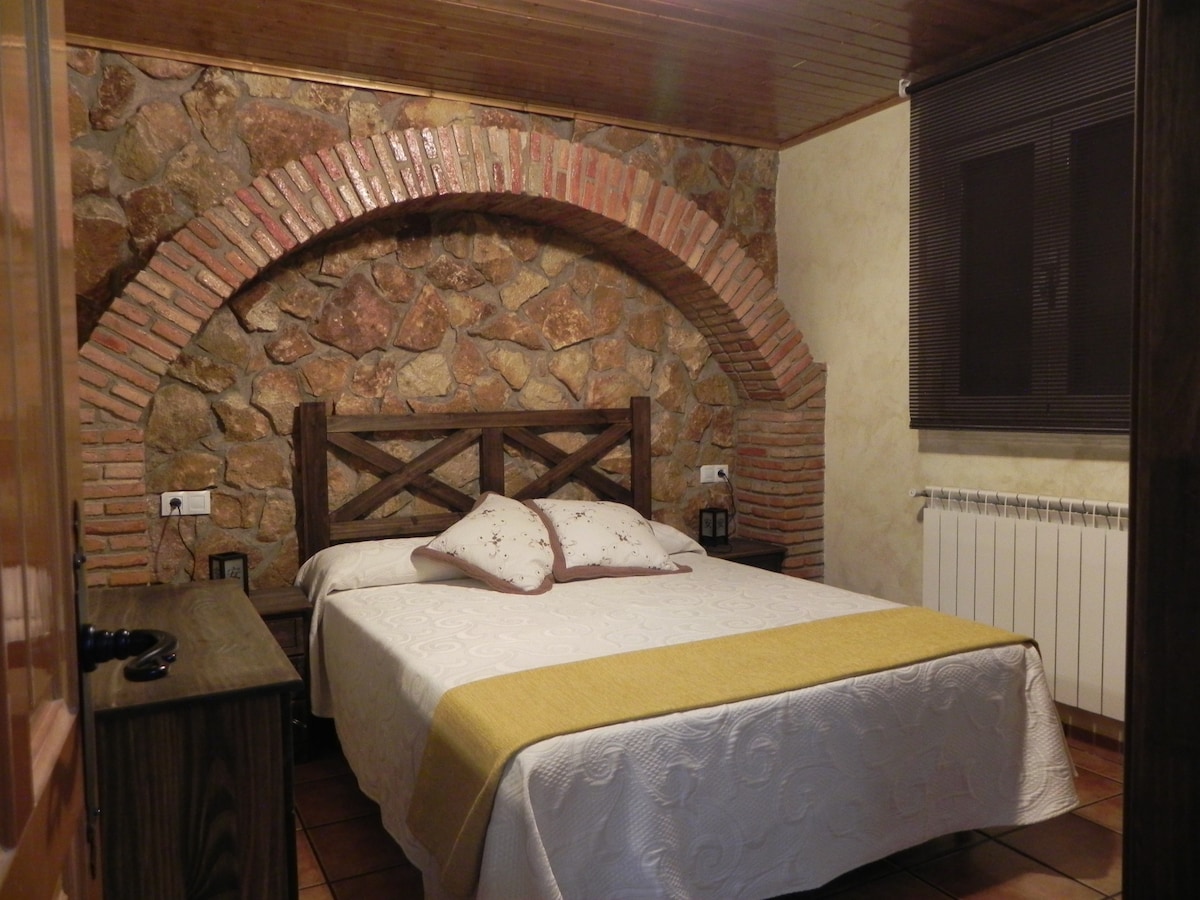 A cozy bedroom features a double bed with light bedding and decorative pillows. A rustic stone wall arches above the bed, complemented by warm wooden details. Two bedside tables are positioned on either side, and a radiator is nearby, enhancing comfort in the room.
