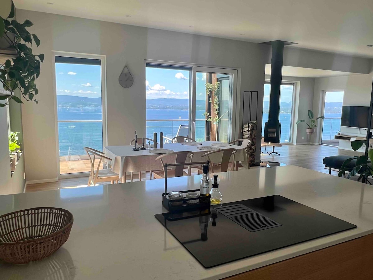The open living area features large windows offering a view of the fjord. A modern kitchen island is positioned in the foreground with simple kitchenware. A dining table is visible, set for meals, while the living area includes a cozy seating arrangement and a fireplace.
