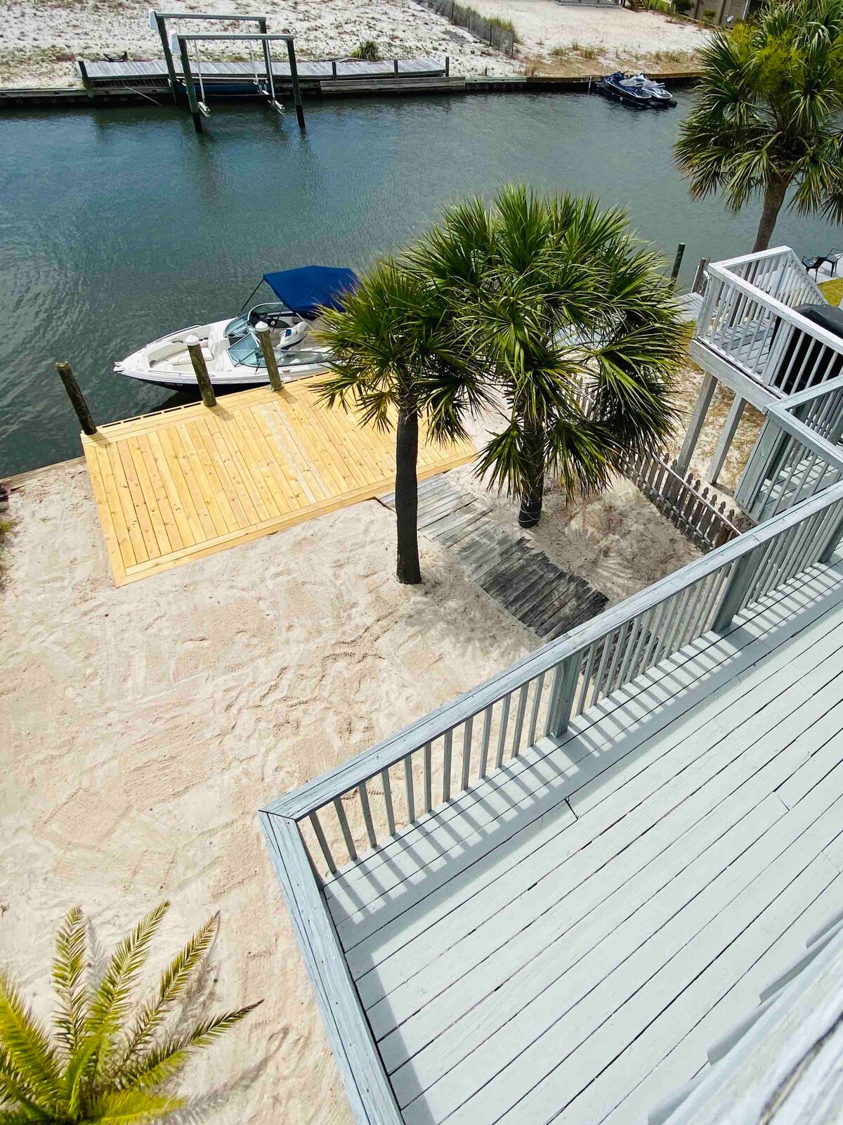 An aerial view of the backyard highlights a sandy area with a wooden deck leading to the water. Palm trees provide shade, and a boat is visible moored nearby, reflecting the waterfront access of the property.