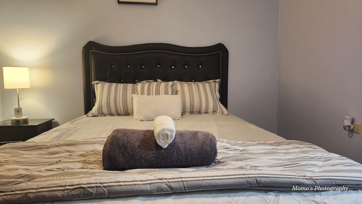 A sleek, upholstered headboard frames a spacious bed adorned with striped linens and decorative pillows. A rolled towel is placed at the foot of the bed, while a bedside lamp emits a soft glow, contributing to the calming atmosphere of the room.