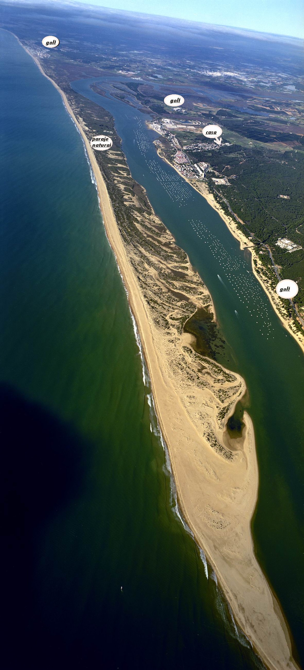 An overhead view of a coastal landscape featuring a sandy peninsula bordered by turquoise waters. The shoreline is marked by gentle waves, while adjacent waterways showcase various boats and lush greenery in the background. The expansive scene captures the serene beauty of the natural environment.