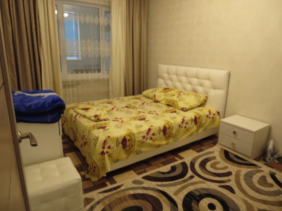 A cozy sleeping area is furnished with a spacious bed covered in a yellow floral-patterned blanket. A nightstand is positioned beside the bed, and a plush seating area is visible. Natural light filters through the window, paired with soft curtains.