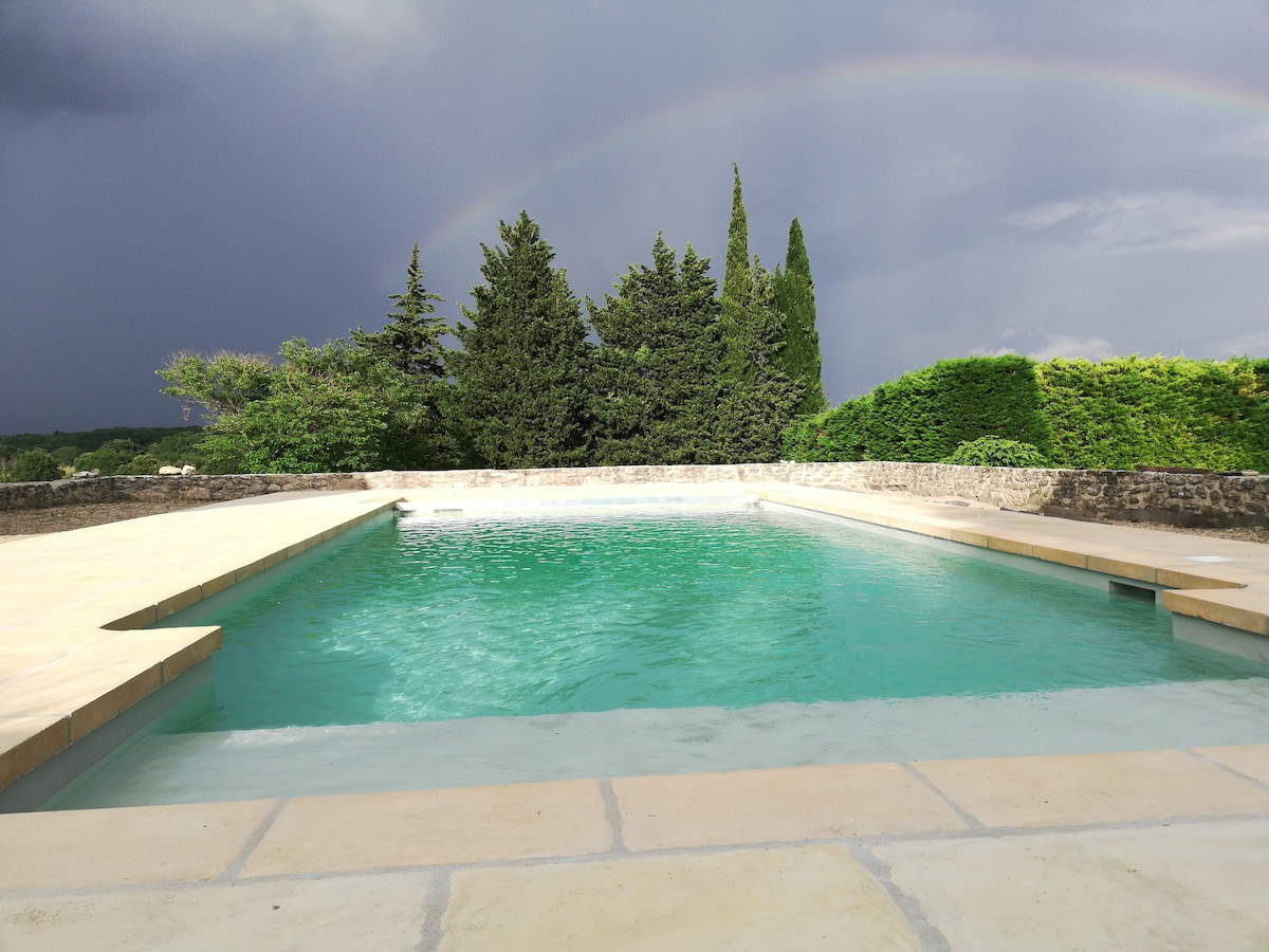 A clear swimming pool is set against a backdrop of lush greenery and a rainbow in the sky. The pool area features stone decking and is surrounded by tall cypress trees, offering a peaceful retreat. The surface of the water reflects the surrounding scenery.