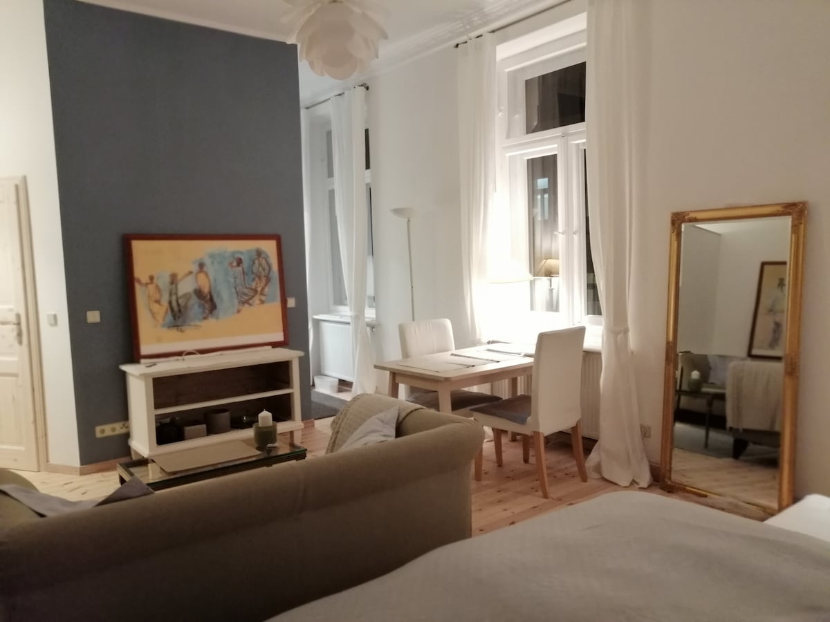 A brightly lit living space features a blend of modern and classic furnishings. A spacious sofa is positioned in front of a large, framed artwork. A dining table with white chairs is visible, along with a full-length mirror reflecting the natural light from the tall windows.
