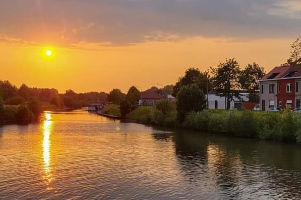 A sunset casts a warm glow over a peaceful waterway, with reflections shimmering on the surface. Lush greenery lines the banks, and residential buildings are visible in the distance, creating a serene riverside atmosphere.