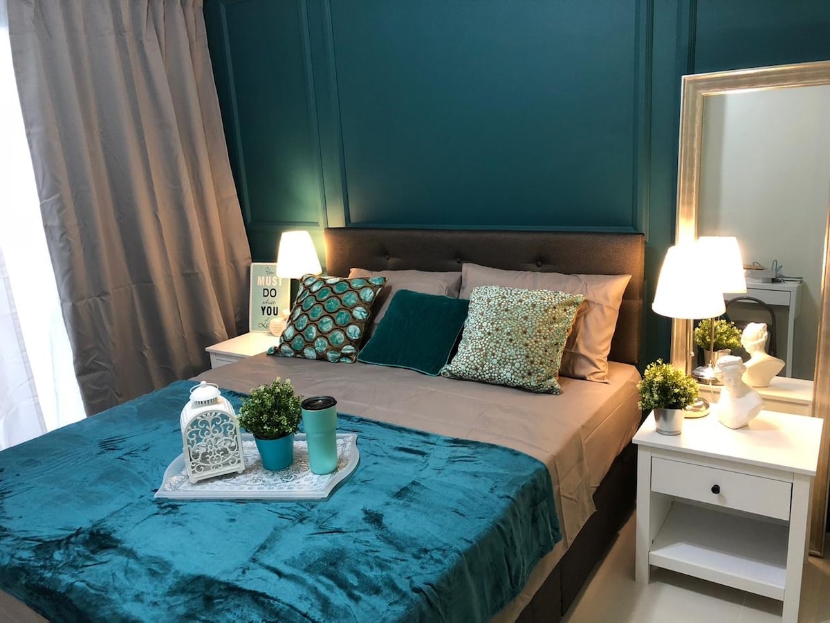 A cozy sleeping area features a spacious bed adorned with teal and gray tones. Two bedside lamps illuminate the scene, complemented by decorative pillows. A small table displays a decorative lantern and potted plants, while a mirror adds depth to the space.