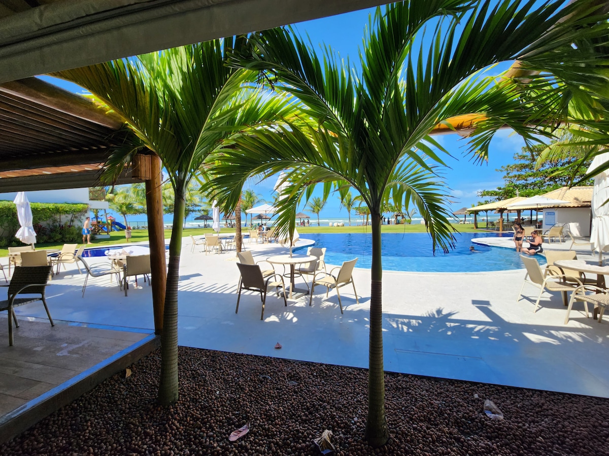 A view of a tranquil pool area framed by lush palm trees. Sun loungers and tables are arranged around the pool, which reflects the blue sky above. The beach is visible in the distance, contributing to a serene coastal ambiance.