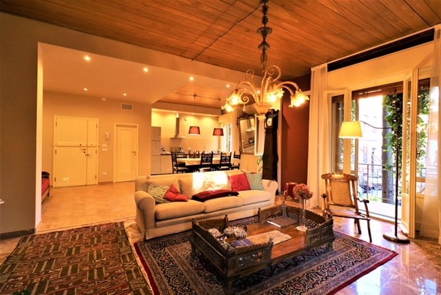 An open-plan living area features a comfortable sofa with colorful pillows and a decorative coffee table. Light from large windows illuminates the space, highlighting the wooden ceiling and tiled floor. A dining table with chairs is seen in the background, along with doors that lead to a terrace.