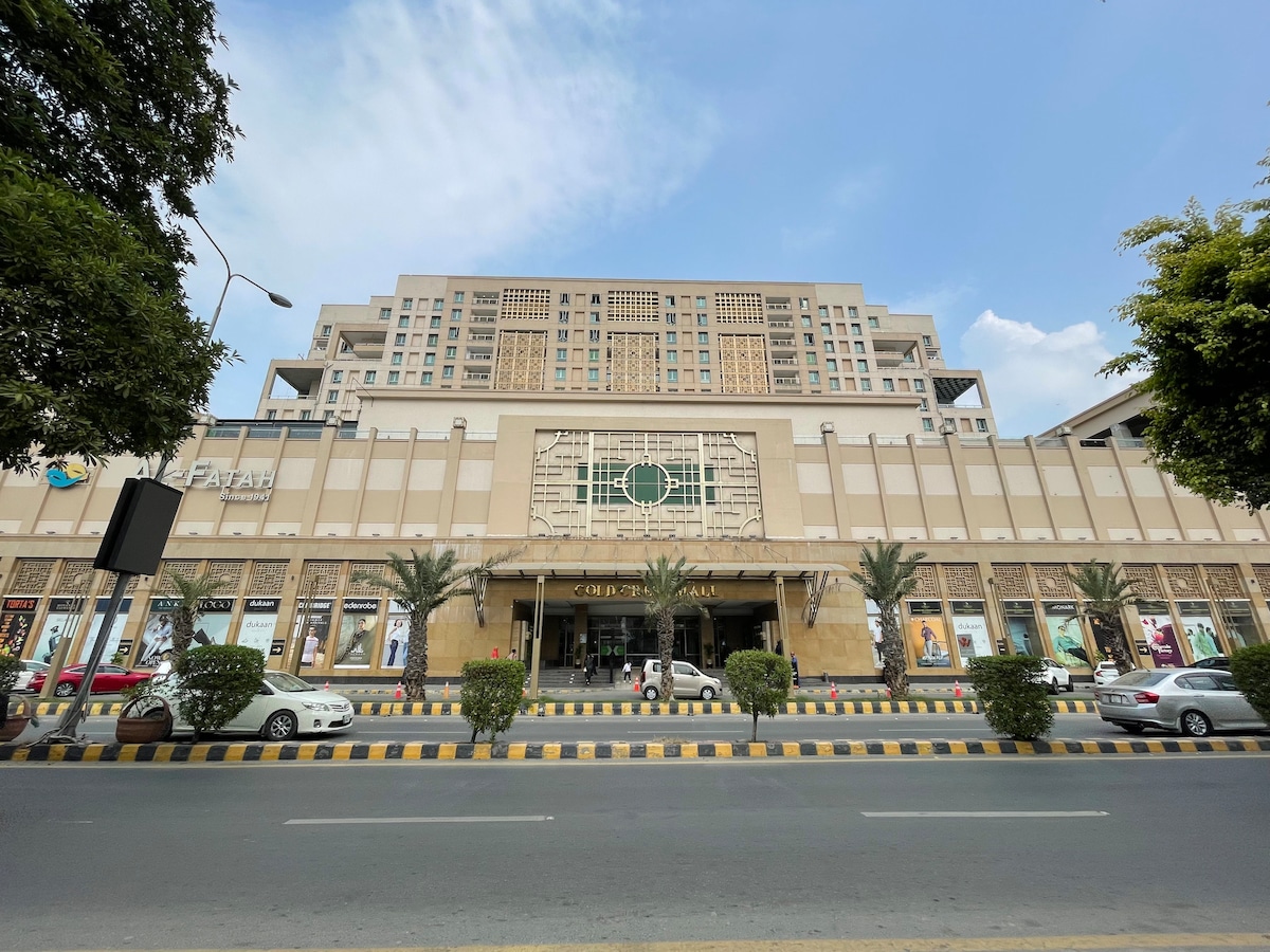 The exterior of Gold Crest Mall & Residency is captured, highlighting its modern architecture and spacious entrance. Palm trees line the walkway, while a variety of vehicles are parked along the street. The sky above is partially cloudy, contributing to the inviting ambiance of the location.