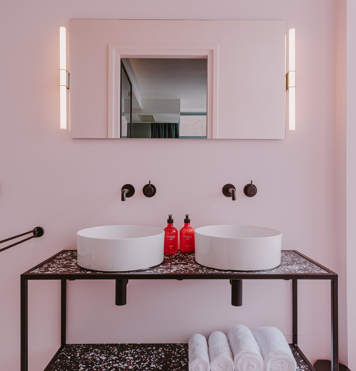 A modern bathroom features a sleek black vanity with two circular sinks atop. The illuminated mirror reflects a hint of the room beyond. Fresh towels are neatly stacked on the side, while vibrant red toiletries add a pop of color against the soft pink walls.