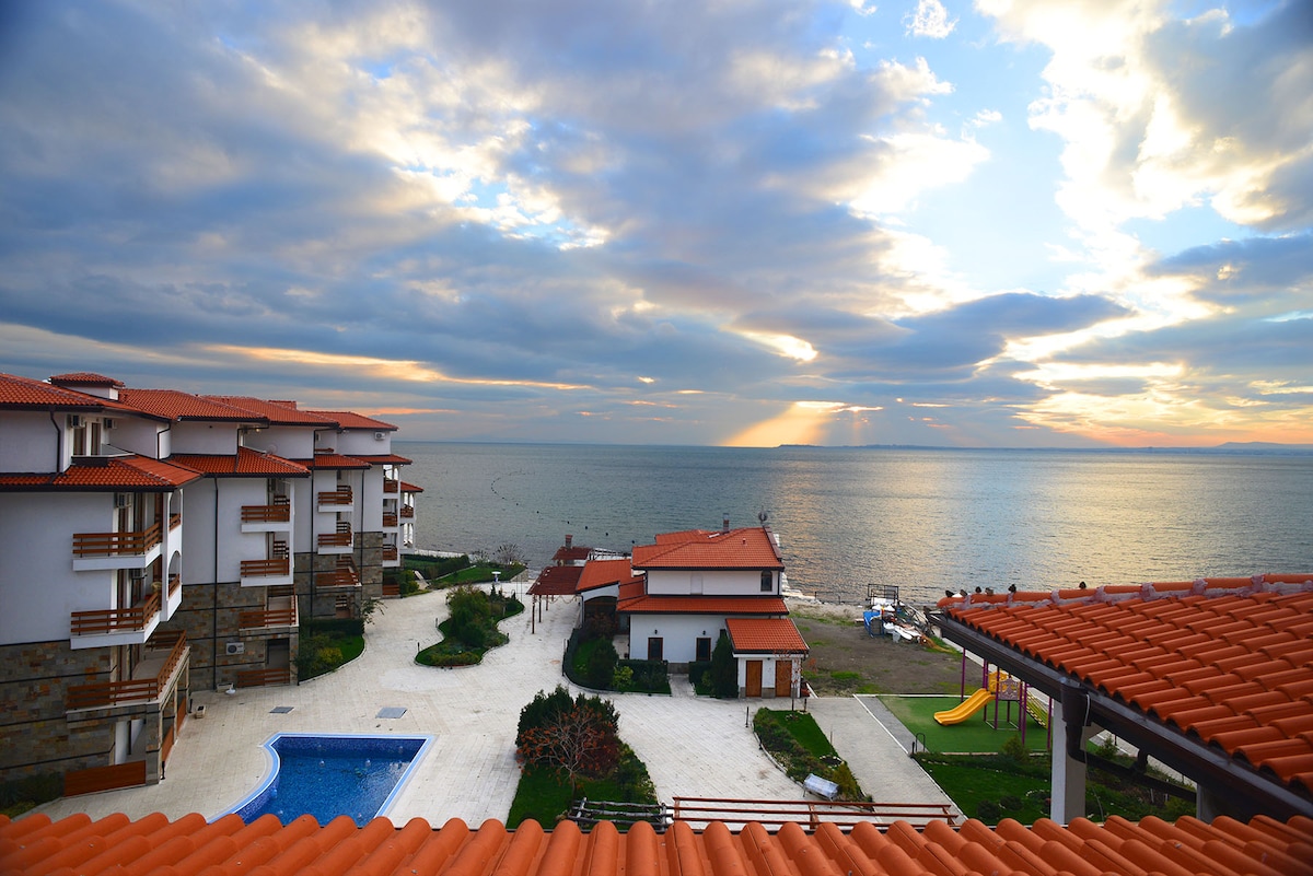 A serene ocean view is presented, showcasing a tranquil blue pool surrounded by well-maintained grounds. Multiple buildings with red-tiled roofs are visible, contrasted against the cloudy sky. The coastline extends toward the horizon, where soft light reflects off the water, creating a peaceful ambiance.