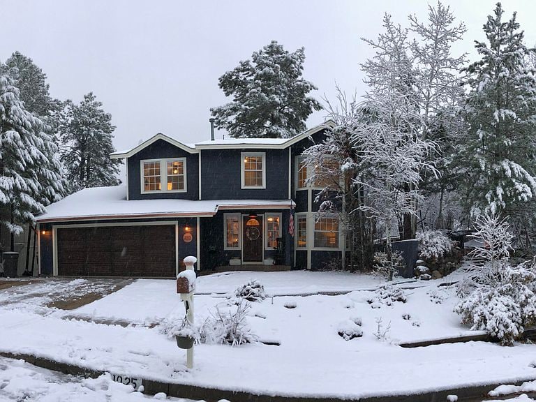 A two-story house is covered in fresh snow, surrounded by tall trees. Warm light glows from the windows, contrasting with the cool tones of the winter scene. The driveway and front walkway are blanketed in white, creating a serene winter landscape.