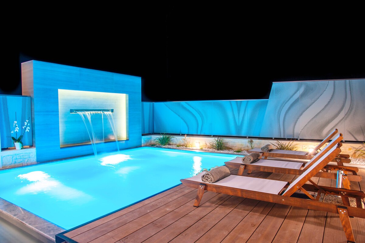 A swimming pool is illuminated in a soothing blue hue, complemented by a waterfall feature that cascades gently into the water. Sun loungers are positioned on a wooden deck nearby, with fresh towels neatly arranged on each lounger, creating an inviting space for relaxation.