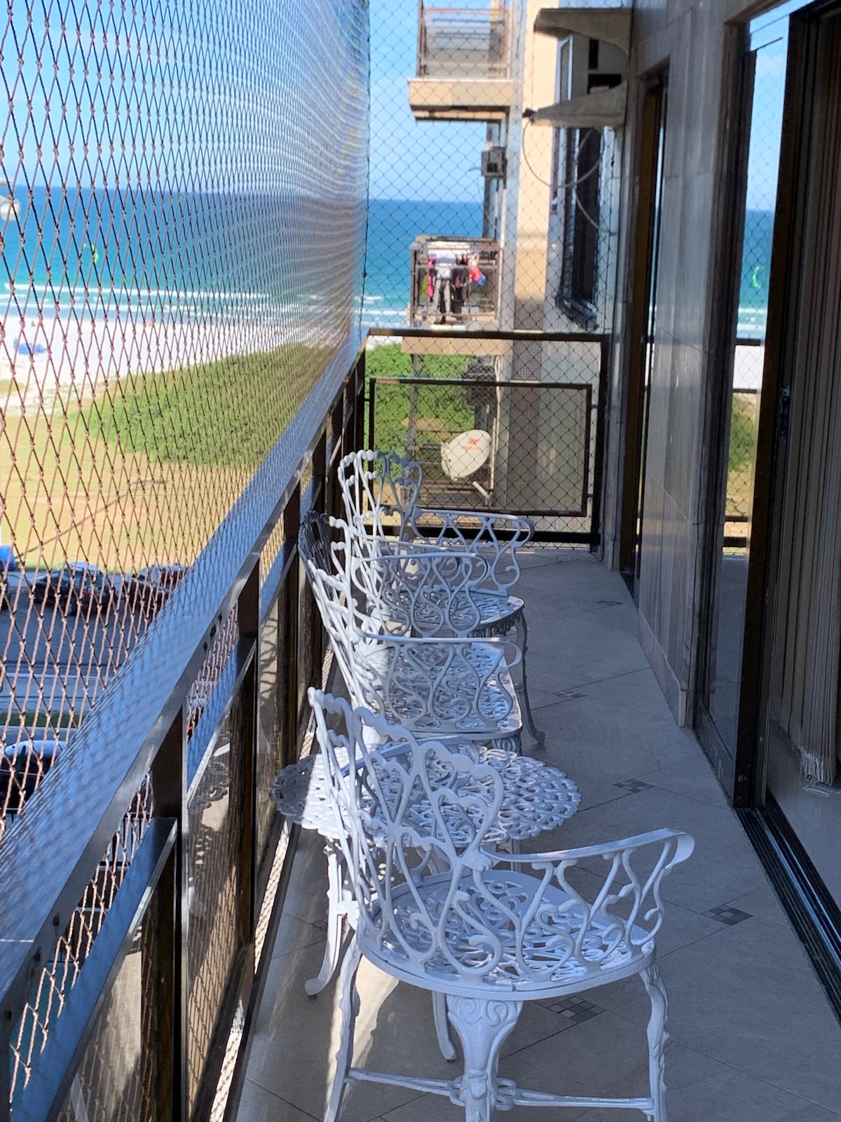 A balcony features a view of the ocean, complemented by a protective netting. Four elegant white chairs surround two small tables, creating a cozy outdoor seating area. The tiled floor adds a polished look, enhancing the space's functionality and charm.