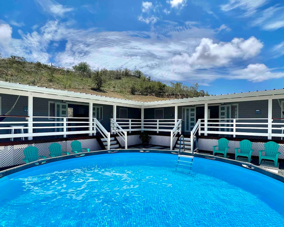 A large swimming pool is surrounded by lounge chairs, offering a serene outdoor space. The pool is framed by a single-story building featuring multiple doors, with a gentle hillside visible in the background under a blue sky adorned with clouds.