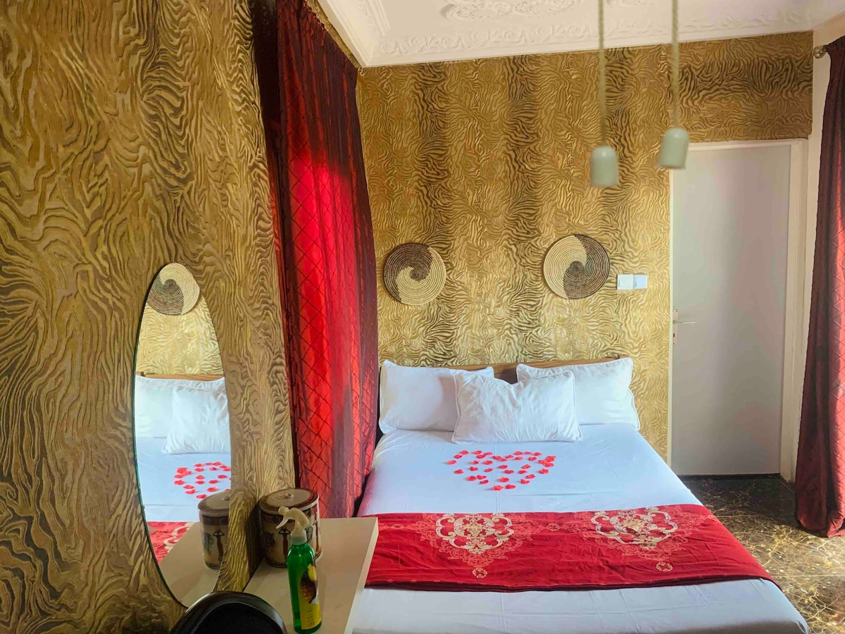 An inviting bedroom features intricate gold-patterned walls, highlighted by deep red curtains. A mirror reflects the spacious bed adorned with decorative red petals and a vibrant red throw. The room is illuminated by soft pendant lighting, creating a warm atmosphere.