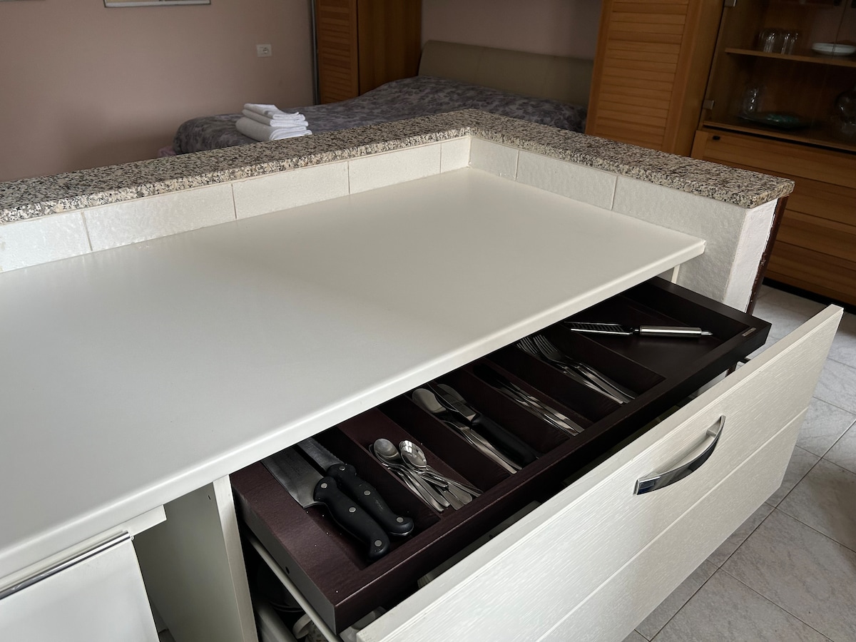 A well-organized kitchen space showcases a white countertop with a partially opened drawer beneath. The drawer contains neatly arranged cutlery, including knives and utensils, with a glimpse of a cozy sleeping area in the background.