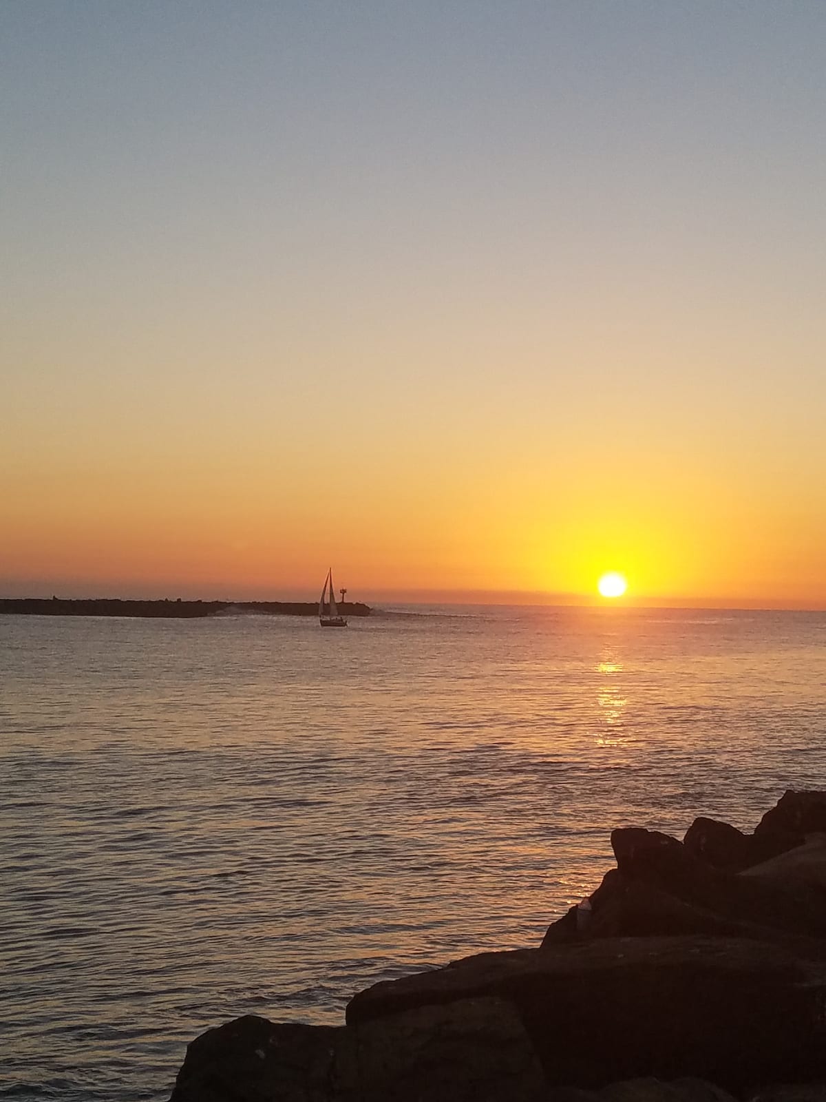 A serene sunset over the water is depicted, with a sailboat gently sailing on the horizon. The sky transitions from warm orange to soft yellow hues, reflecting on the calm surface of the bay.