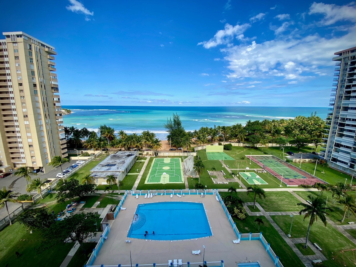 A panoramic view captures the ocean alongside well-maintained recreational facilities. Below, a swimming pool is visible, with adjacent courts for basketball and racquetball. Lush greenery and palm trees frame the scene, while the distant coastline offers a glimpse of the beach and blue waters.