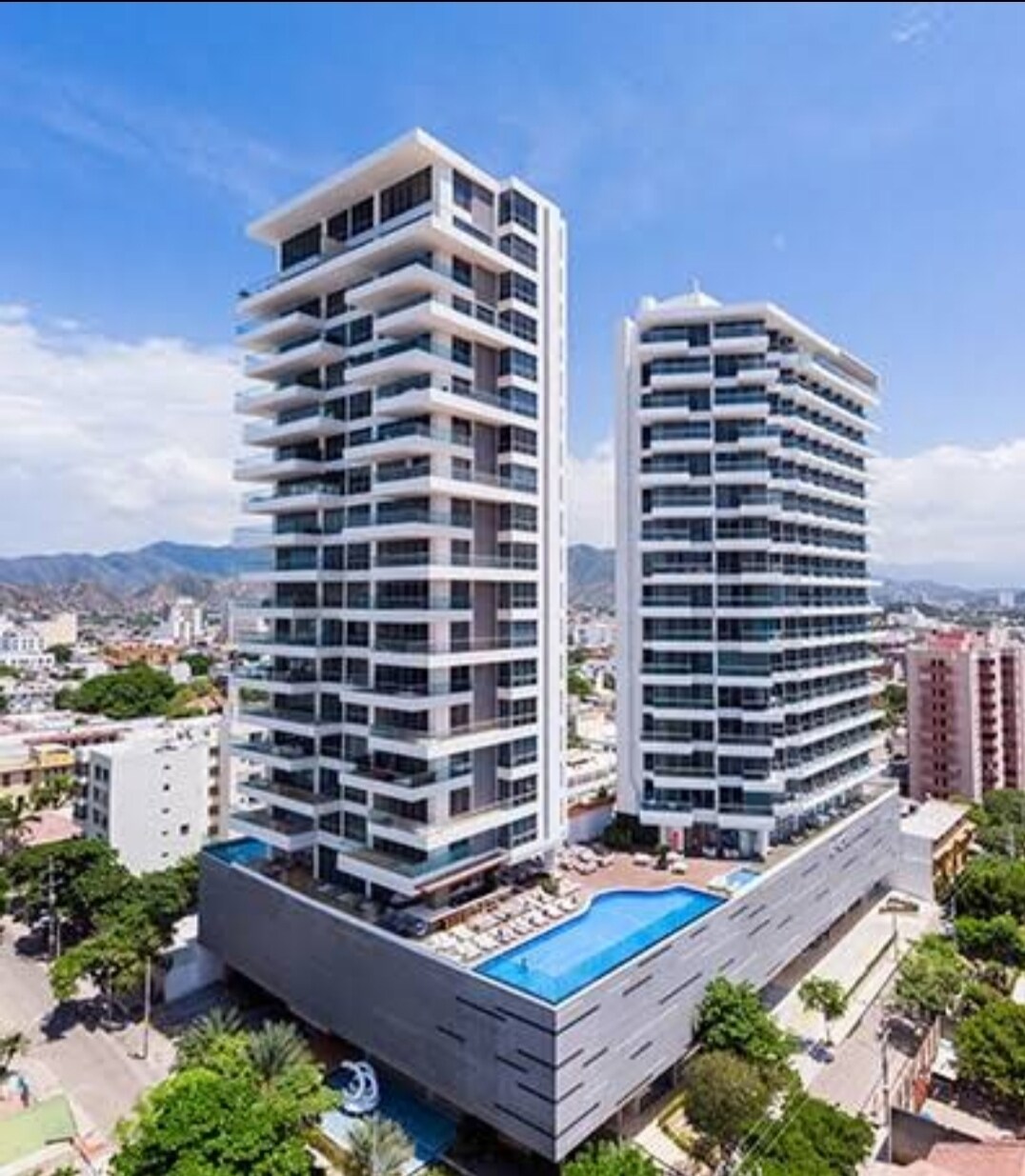 A modern high-rise building complex is presented from an aerial view, showcasing two towers featuring expansive balconies and a shared swimming pool. Lush greenery surrounds the base, while the cityscape and mountains serve as a backdrop, creating a harmonious blend of urban and natural elements.