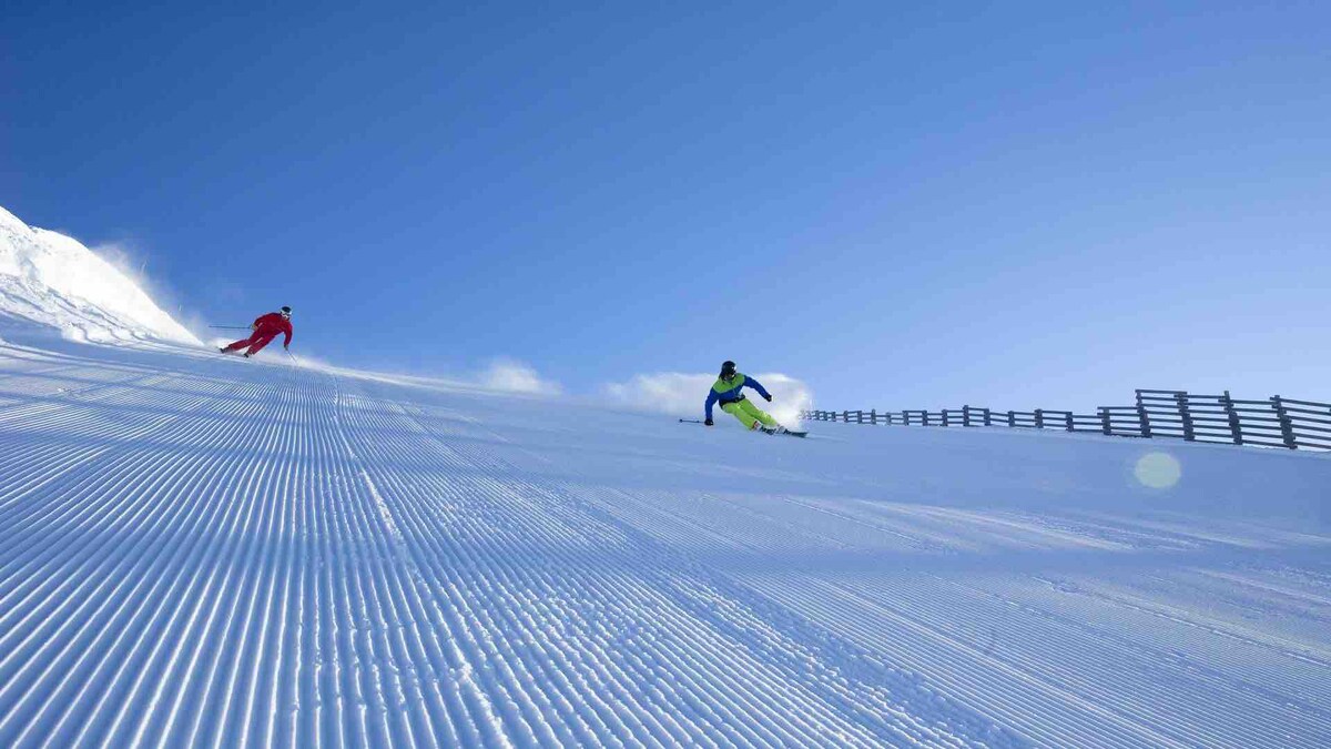 A clear blue sky serves as the backdrop for two skiers navigating freshly groomed slopes. The pristine snow is highlighted by parallel lines created by the grooming machinery. A wooden fence runs along the side of the slope, enhancing the serene winter landscape.