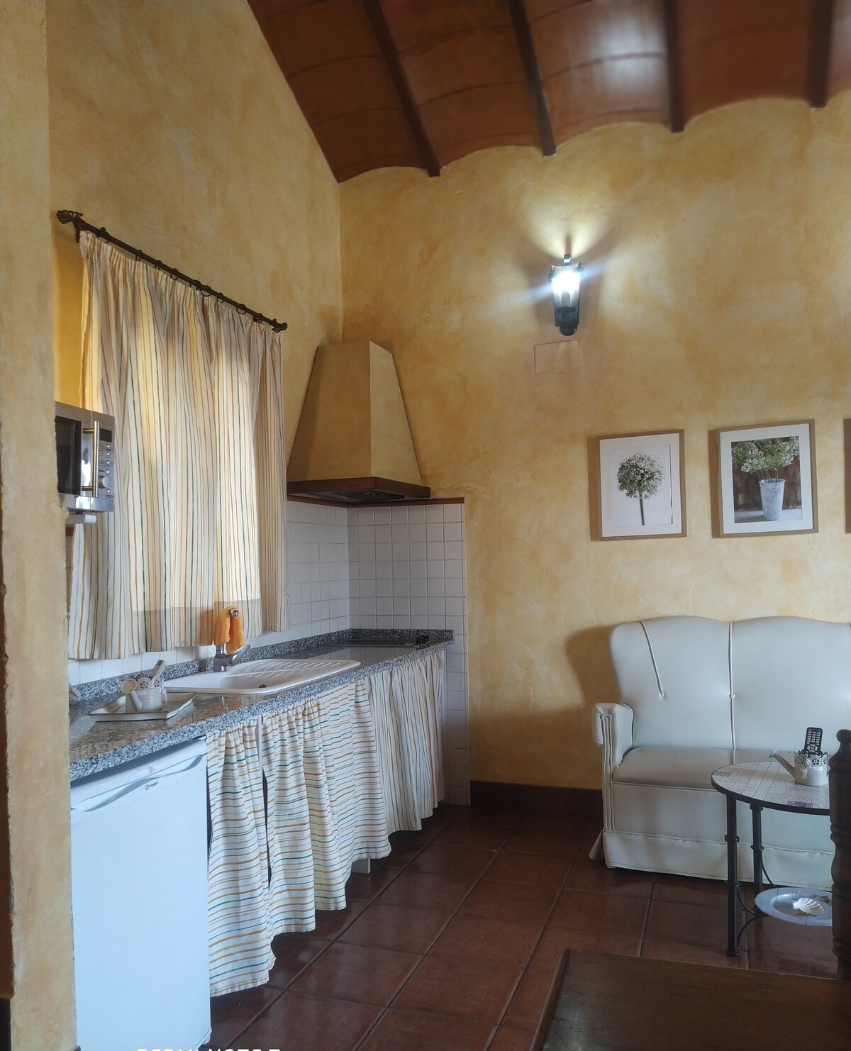 A compact kitchenette is equipped with a refrigerator, microwave, and a granite countertop. Soft yellow walls add warmth to the space, complemented by a white sofa and a small side table. Light-filtering curtains drape over the window, enhancing the cozy atmosphere.